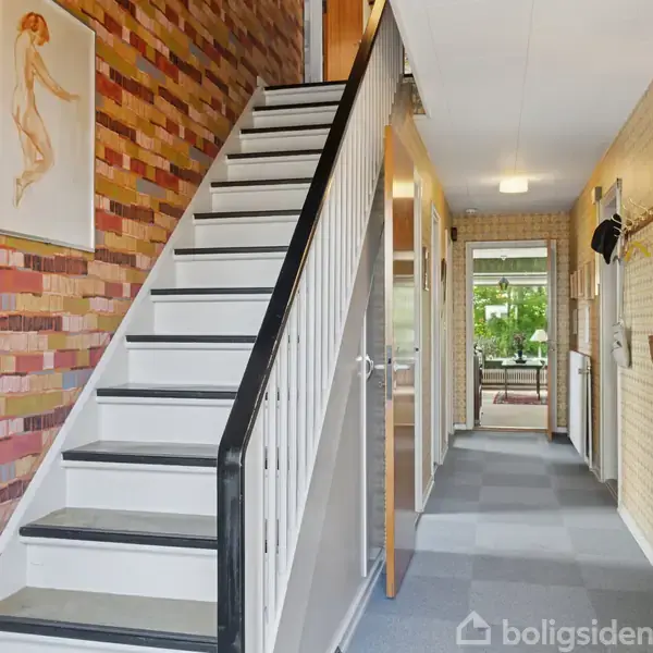 Trappe med sort håndliste stiger i en gang med murstensmønstrede tapeter. Gangen leder til en stue med store vinduer og grønne planter udenfor. Billeder hænger på væggen.