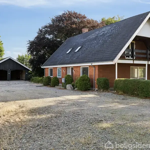 Et rødt murstenshus med sort tag ligger ved siden af en grusopkørsel. Et sort skur står i baggrunden omgivet af træer og buske.