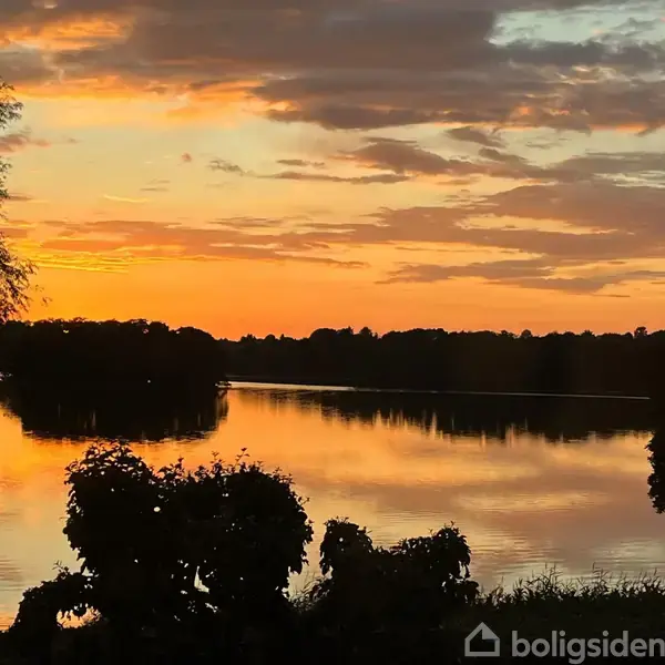 En sø reflekterer en farverig solnedgang, omgivet af silhuetter af træer. Den fredfyldte scene skaber en atmosfære af ro og skønhed.
