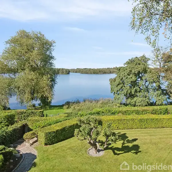 En velholdt have med klippede buske grænser op til en sø omgivet af træer under en klar himmel.