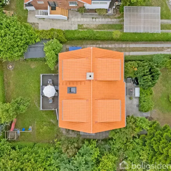 Et hus med et orange tag set ovenfra, omgivet af en grøn have. En terrasse med en paraply ligger ved siden af huset. Der er stier og hække rundt omkring.