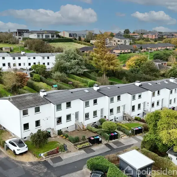 Rækkehuse i hvid facade står roligt ved siden af hinanden i et boligkvarter. Biler er parkeret foran, omkranset af små haver. Baggrund indeholder flere huse og grønne områder.