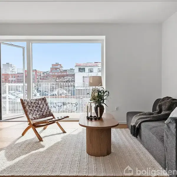 En stue møbleret med en sort sofa og en træstol står i lys fra de store vinduer. En grøn plante står ved siden af, mens et træbord er centralt placeret.