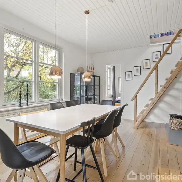 Et spisebord med seks stole står midt i et lyst rum med store vinduer. En trappe fører op til en hems, og der er billeder på væggen.