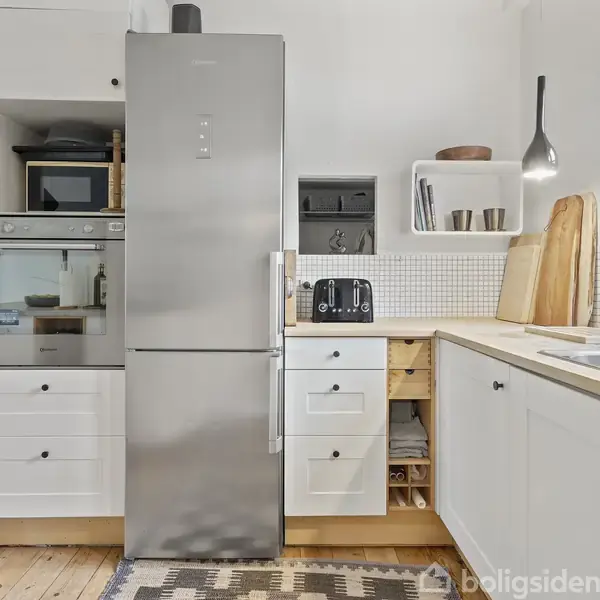 Køleskab i rustfrit stål står ved siden af hvidt køkkenbord. Kombinationen af tæpper på trægulv, brødrister, træskærebrætter og køkkenskabe skaber en moderne køkkenstemning.