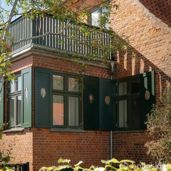 Et rødstenshjørnehus med grønne skodder åben ved vinduerne. Skodderne har små dekorationer. En balkon og trægrene kan ses øverst og til venstre.