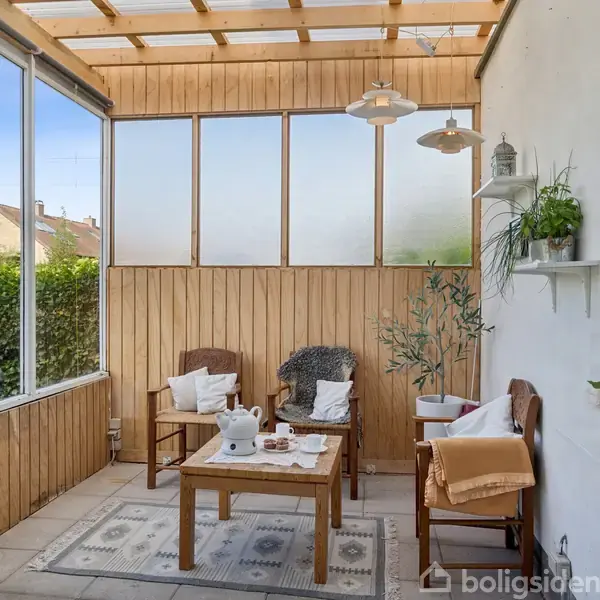En hyggelig veranda med trævægge og loft, indeholdende to træstole og et bord med kaffe- og testel. Omgivelserne består af planter, hylder og naturligt lysindfald fra store vinduer.