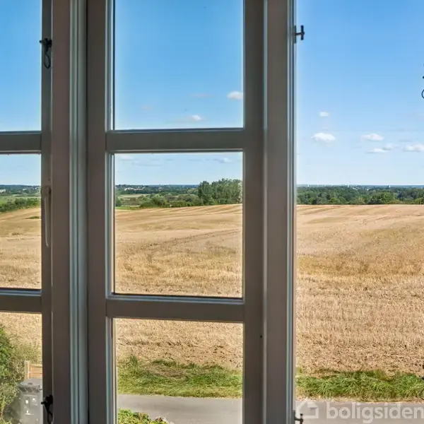 Vindue åbner mod en gylden kornmark. Marken strækker sig til horisonten, omgivet af grønne træer under en klar, blå himmel. En vej ses i forgrunden, tæt på vinduet.