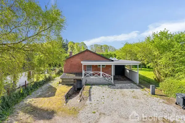 Toilet står ved siden af en træstol under et lille kvadratisk vindue i et badeværelse. Et stort spejl og skabsarrangement fylder væggen på højre side. Gulvet er hvidt med fliser.