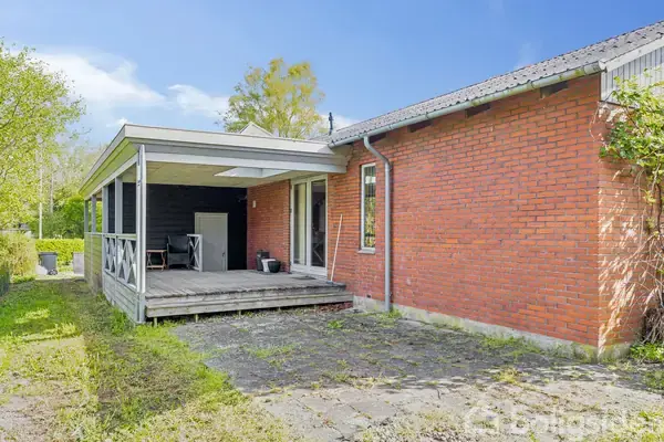 Toilet og bruser står i enkelt badeværelse med flisegulv og træbeklædt loft. Toiletrullen hænger ved siden af toilettet, og vindue med dekorative genstande åbner til lys udenfor.