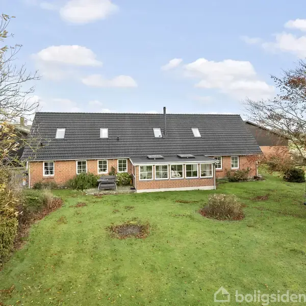 Rødt murstenshus med sort tag og flere ovenlysvinduer. Foran huset er en grøn have med buske og træer. Himlen er delvist skyet.