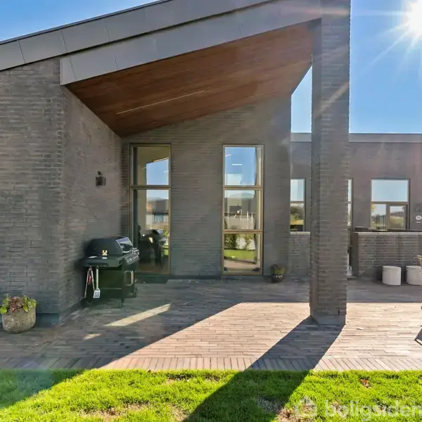 Bygning har fladt tag i moderne stil. Terrasse med grill og havemøbler, omgivet af græsplæne under klar himmel i solskin.