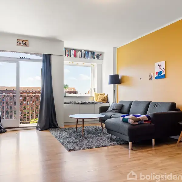 En stue med en grå sofa og sofabord står på et tæppe foran en gul væg. En stor glasdør fører til en balkon med udsigt til en bygning.