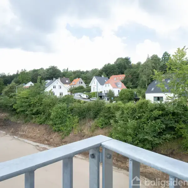 Balkon med metalgelænder, omkranset af grønne buske. I baggrunden ses flere huse med forskellige tagfarver, omgivet af træer under en delvist overskyet himmel.