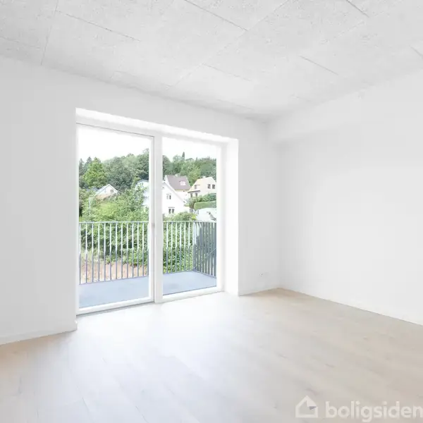 Et tomt værelse med lyse trægulve og hvidmalede vægge. En stor glasdør fører til en balkon med udsigt over grønne omgivelser og et par huse i baggrunden.