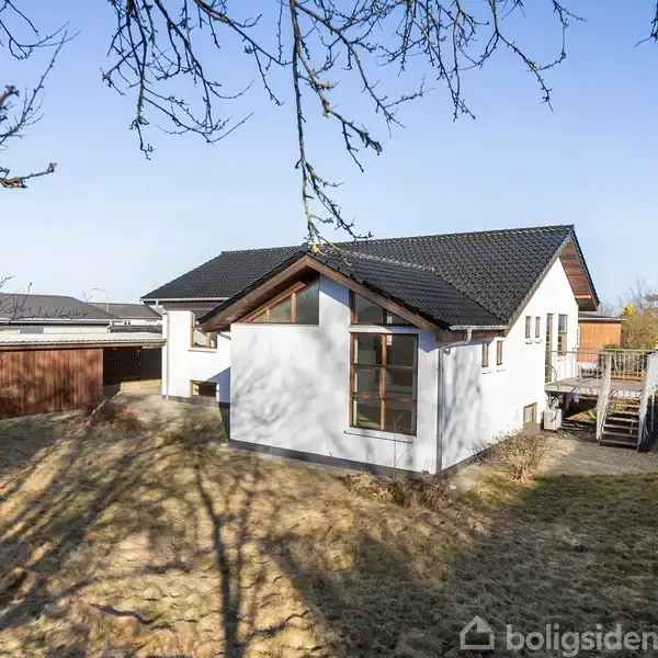 Hvidt hus med sort tag står stille i en have med græsplæne og omgivende træer. Terrasse med trin til venstre. Skyfri himmel i baggrunden.