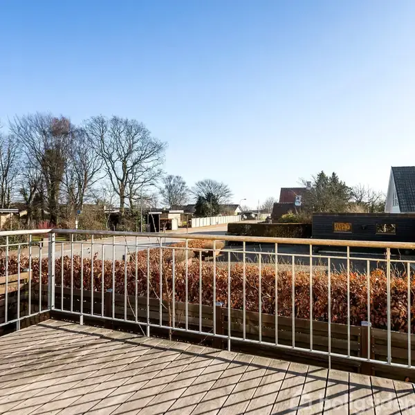 En træterrasse med metalgelænder står i forgrunden. Buske flankerer terrassen, mens villaer og træer kan ses i det omkringliggende bymiljø under en klar blå himmel.