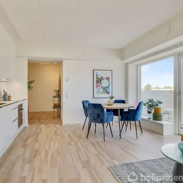 Et køkkenalrum med moderne, minimalistisk indretning, hvor et rundt spisebord med blå stole står til højre. Store vinduer giver lys og udsigt. Planter pynter rummet.