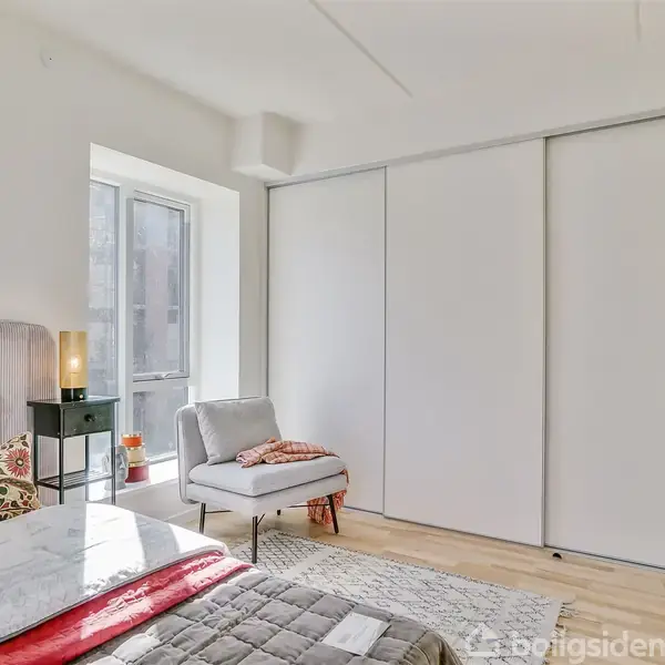 Soveværelse med en seng i forgrunden, en lænestol ved vinduet og en stor skydedørsgarderobe langs væggen. Rummet er lyst med trælaminatgulv. Ingen tekst til stede.