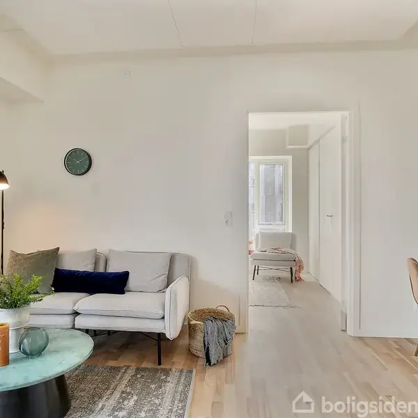 Stue med en lysegrå sofa, ved siden af en gulvlampe og et lille bord med planter. En åben dør viser et tilstødende rum. En stol ved skrivebordet til højre.