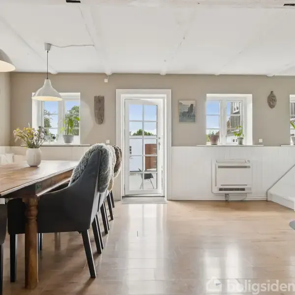 Spisebord med stole står i en lys stue, hvor en brændeovn er i brug. Omkring er der vinduer og en dør, der fører ud til en terrasse.