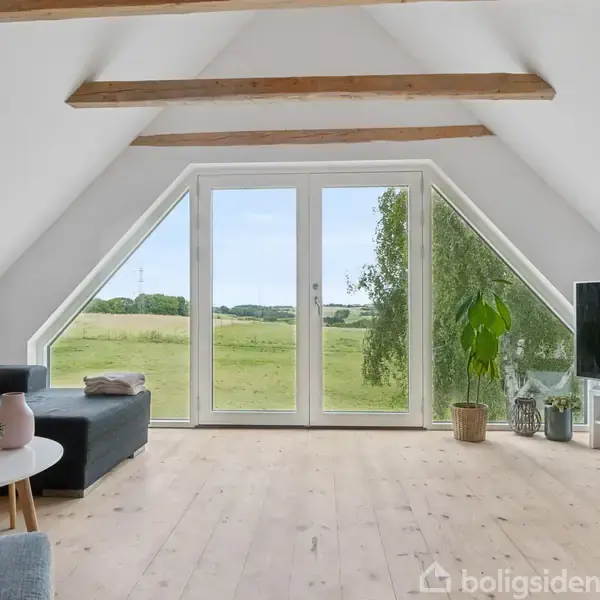 Stue med grå sofa og TV viser udsigt til en mark gennem store vinduer. Trægulv og fritliggende loftsbjælker skaber en lys og åben atmosfære.