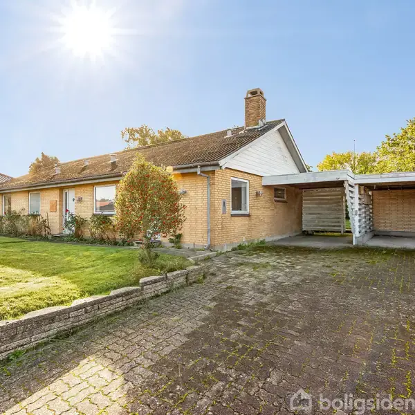 Murstenshus med skråtag står under en klar himmel, omgivet af en velplejet forhave og en belagt indkørsel med tilhørende carport. Solen skinner klart i baggrunden.