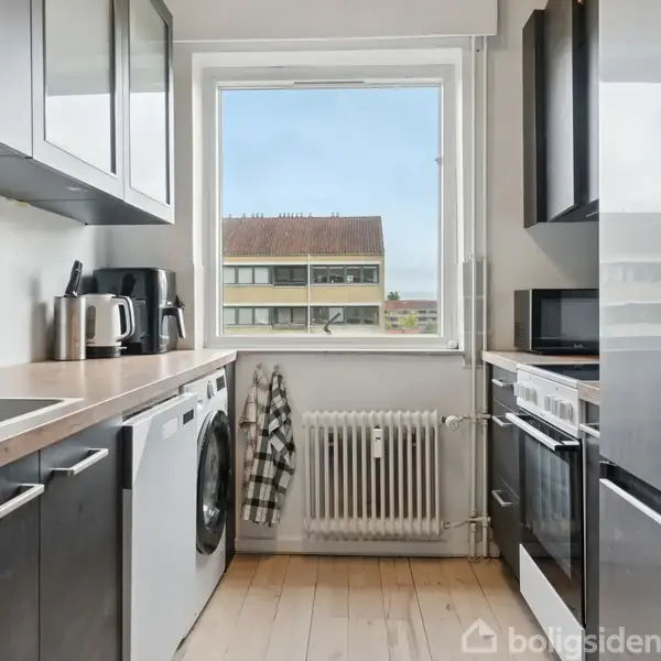 Køkken med sorte skabe har vask og opvaskemaskine til venstre, ovn og mikroovn til højre, mug og elkedel på bordpladen. Ud af vinduet ses en bygning i baggrunden.