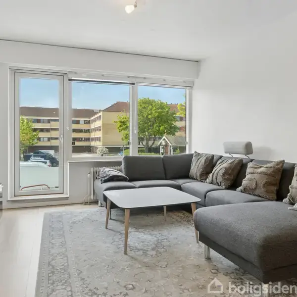 En grå hjørnesofa står i en stue med et sofabord foran. Et stort vindue viser en udsigt til træer og bygninger udenfor. En tv-bænk med tv er til venstre.