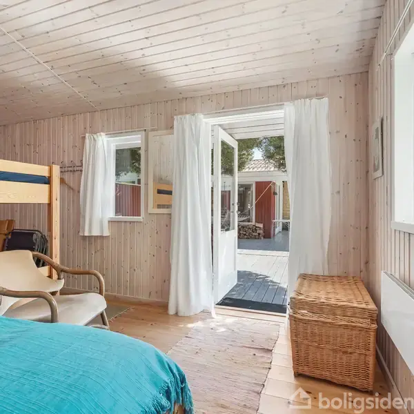 Et værelse med køjeseng står ved siden af en lænestol. Dobbelt døre åbner ud mod en terrasse. Et kurvemøbel og lyse trævægge er synlige.