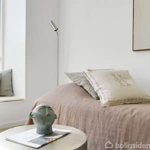 Soveværelse med en seng dækket af beige sengetæppe og puder, stående ved siden af et rundt bord med en keramisk figur. Til venstre ses en vindueskarm med hynder.
