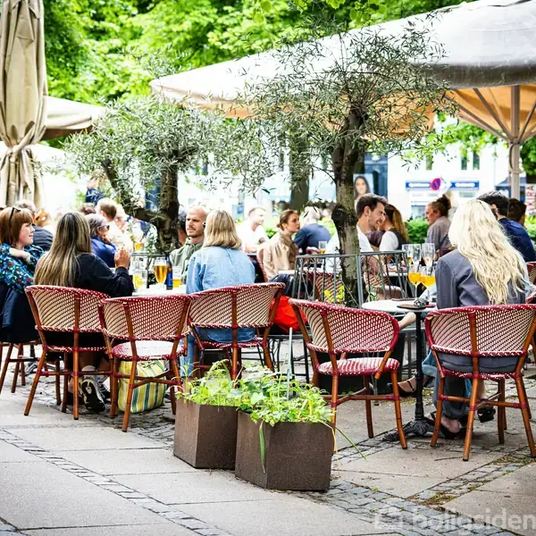 Folk sidder og spiser og drikker ved cafe borde under parasoller. Træer og planter omgiver den udendørs terrasse, der skaber en hyggelig atmosfære.