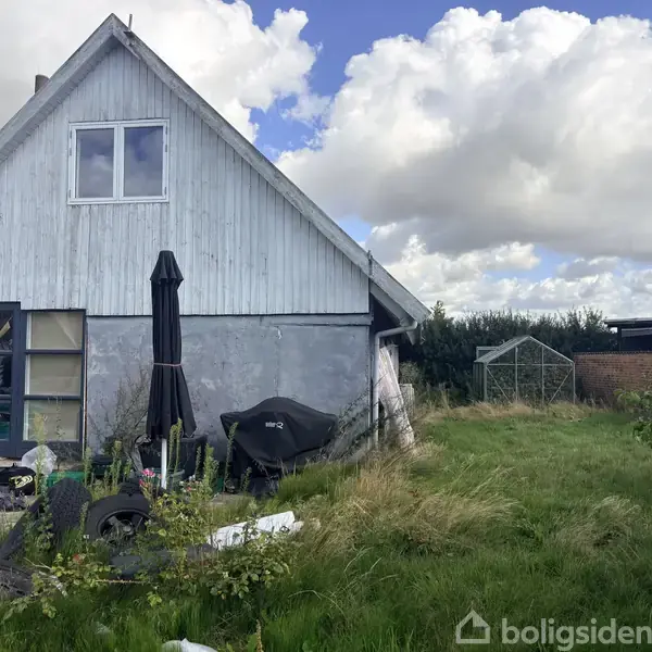 Et hus med gråt tag står ved siden af en rodet have, hvor græsset er langt. En terrasseparasol og en grill er tæt på huset. Himmel med skyer er i baggrunden.