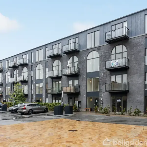 Et moderne lejlighedskompleks med mørke mursten og glasaltaner står i et regnvådt, bymæssigt område. Flere biler er parkeret foran bygningen, der blander indgangspartier med buede vinduer.
