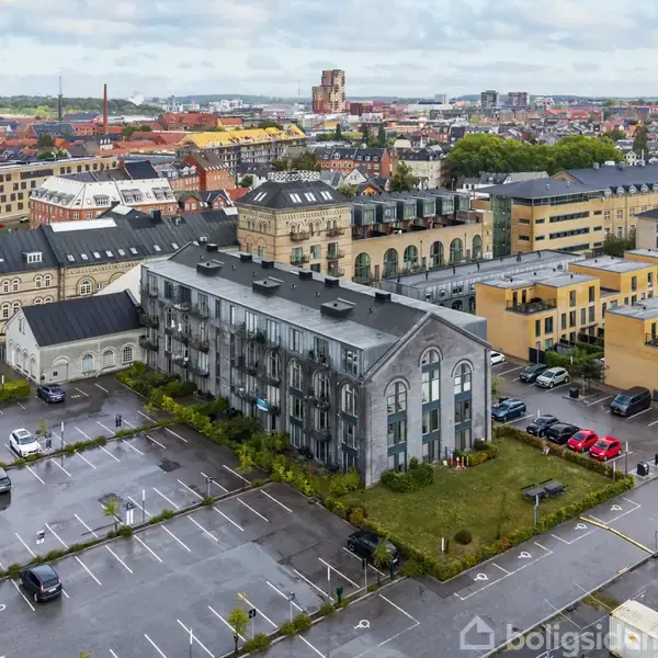 En række bygninger omgiver en central, ældre bygning med moderne glasdetaljer, beliggende ved en parkeringsplads med få biler. Byen strækker sig i horisonten under en overskyet himmel.
