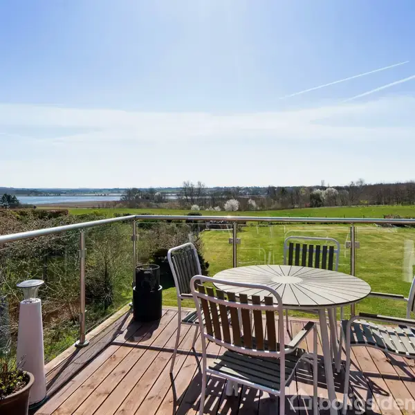 Rundt havebord med stole står på en træterrasse. Terrassen har glasgelænder, og i baggrunden ses grønne marker og en sø under en klar, blå himmel.