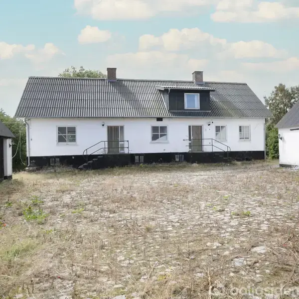 Hvidt landhus står stille mellem to lader på en brostensgårdsplads under en skyet himmel. Trappen fører op til hoveddøren, og lave buske vokser langs væggene.