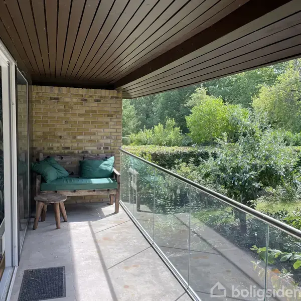 En balkon med glasrækværk og træloft har en bænk med grønne puder og en lille skammel. Den omgivende have indeholder tæt grøn bevoksning og murede vægge.
