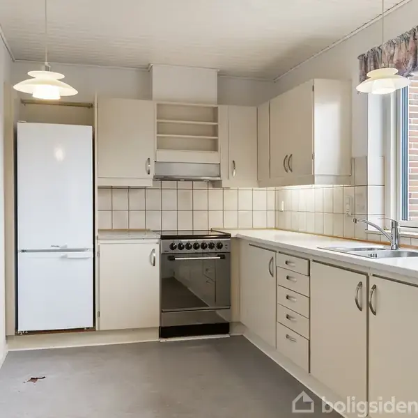 Køkken med hvide skabe og skuffer, køleskab, ovn og håndvask; to loftslamper hænger over grå gulvplader. Flisebelagt væg ved bordplade, vindue til højre med udsigt til mursten.