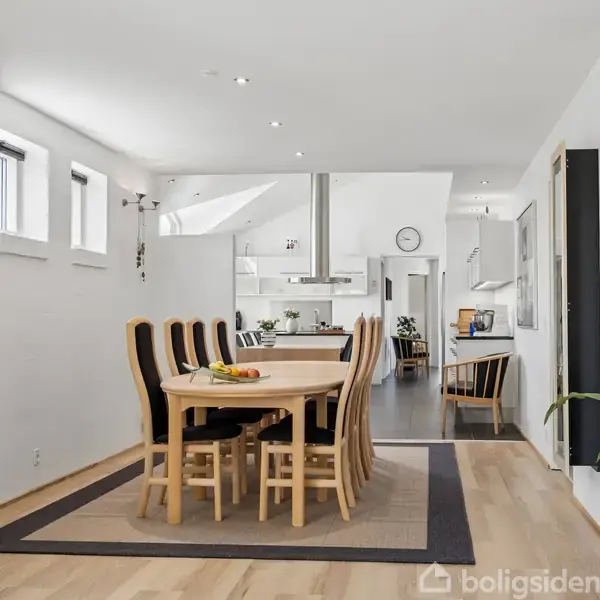 Spisebord med seks stole står på et tæppe i en moderne, lys spisestue med planter. I baggrunden ses et åbent køkken med metalemhætte og vægur.