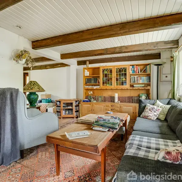 Stue med en grå sofa og en lænestol, der omkranser et træbord. Hylder med bøger og udsmykket skænk i baggrunden. Hvidmalet loft med synlige træbjælker. Ingen tekst fundet.