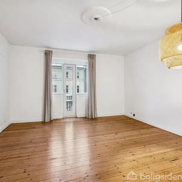 Et tomt værelse med trægulv indeholder en hængende lampe. Store vinduer med gardiner viser udsigt til bygninger udenfor.