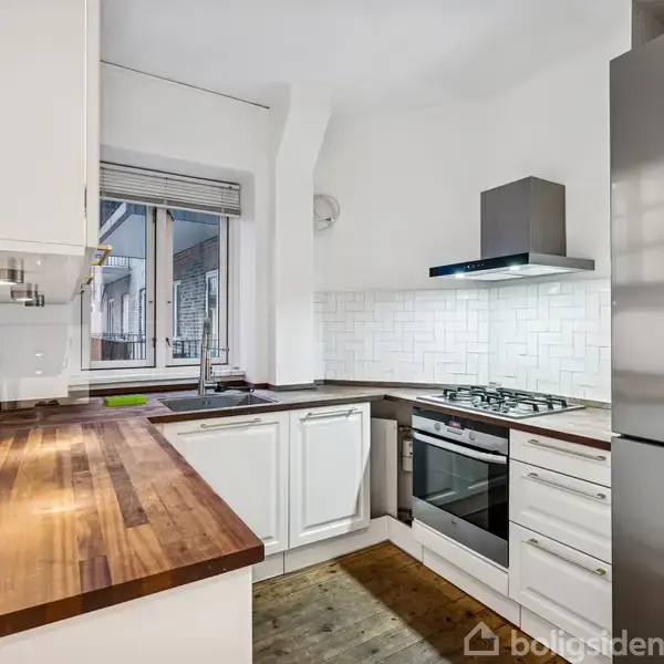 Et moderne køkken med hvide skabe og træbordplade. Et gaskomfur med ovn under emhætte. Et køleskab står til højre. Et vindue giver lys og udsigt udenfor.