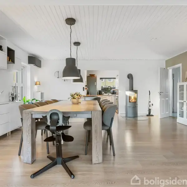 Et moderne spisestue med et stort træbord omgivet af stole. En brændeovn står i hjørnet. Rummet har hvide vægge, trægulv og indbyggede hylder med dekorative genstande.