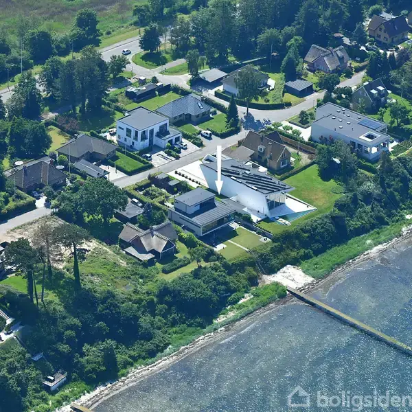 Et moderne hus med hvidt, geometrisk design ligger blandt andre huse i et grønt boligområde ved en kystlinje. Stien og havet er synlige i nærheden.