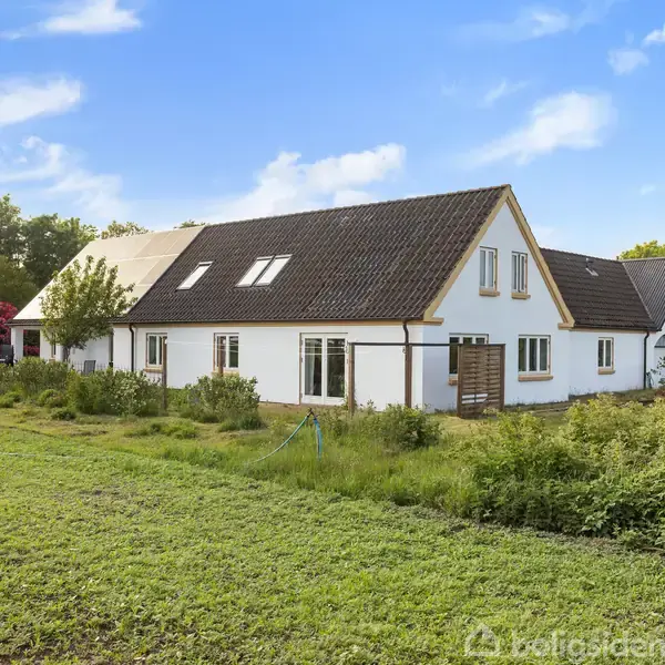 Et hvidt, langstrakt hus med mørkt tag står blandt grønne planter og buske. Baggrunden består af træer under en blå, skyet himmel.