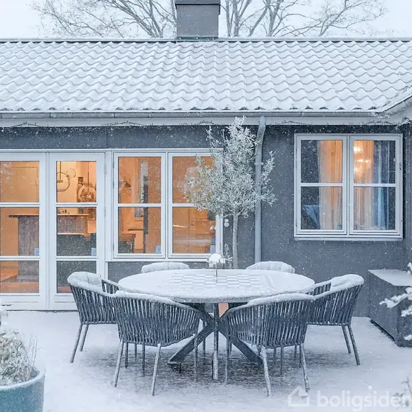 Et rundt havebord med stole er dækket af sne. Det står på en terrasse foran et hus med store vinduer, hvor varm belysning lyser op indefra. Et stort træ står i baggrunden.