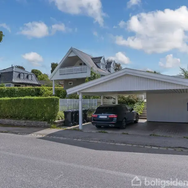 Hus med hvid facade og grå tag, terrasse med liggestole, omkranset af grønne buske og græsplæne, i solrigt vejr under blå himmel.