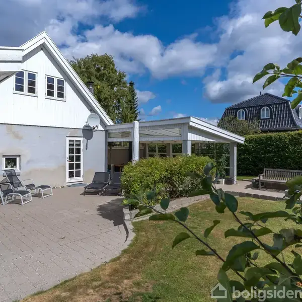 Et hvidt hus med skrånende tag står på en velplejet græsplæne. Ved siden af er en overdækket terrasse, omgivet af buske og naboer tæt på.