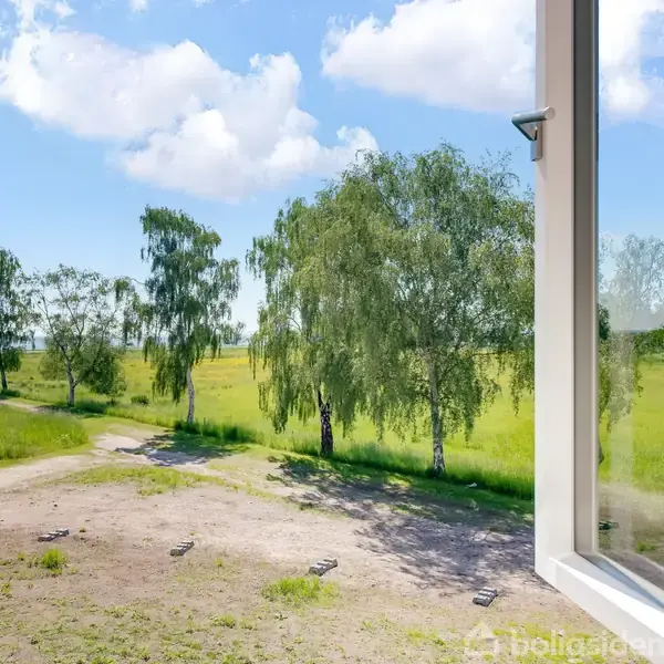 Et åbent vindue vender ud mod en frodig mark. Træer står langs en grusvej, mens blå himmel og hvide skyer præger baggrunden.